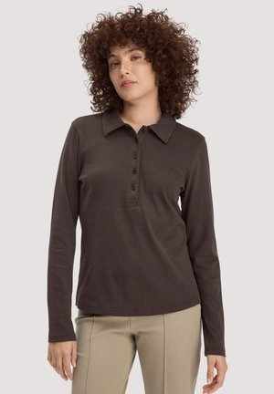 Junge Frau mit lockigem Haar, die ein dunkelbraunes, langärmeliges Hemd mit Kragen und beigen Hosen trägt, vor einem schlichten Hintergrund stehend.