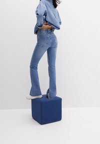 Blue denim flared jeans with a high waist and distressed hem, paired with a matching denim jacket, standing on a blue cube. White sneakers visible.