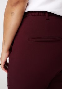 Bordeaux elastische taillebandbroek met een gladde textuur, voorzien van een enkele achterzak en een nauwsluitende pasvorm, tentoon gesteld naast een blote arm.