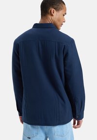 marineblauwe button-up overhemd met lange mouwen, gemaakt van zachte stof, met een kraag en een enkele achterkant yoke-detail, gedragen over lichte denim.