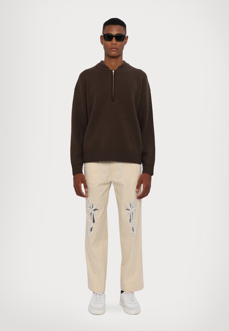 Maglione marrone a maglia con collo a mezza zip, pantaloni crema con intricate ricami neri e argentati, e sneakers bianche.
