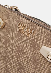 Brązowa torebka monogramowa z złotymi akcentami i teksturowanym materiałem, na którym wyraźnie widnieje logo "GUESS EST 1981".