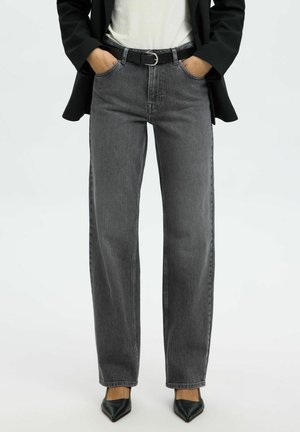 Person som har på seg høytlivs, rette grå jeans med svart belte, svarte spisse hæler, hvit topp og svart blazer med hendene i lommene.