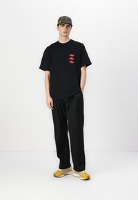Svart oversized t-skjorte med røde grafikkdetaljer, kombinert med løse svarte bukser og gule sneakers med grå detaljer, supplert med en kamuflasje-cap.