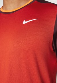 Nike urheilupaita kirkkaan punaisena, mustilla hihoilla, tekstuurilla varustetusta kankaasta ja oranssilla kauluksessa. Rinnassa on valkoinen logo.