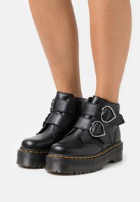 Black leather ankle boots with double heart-shaped buckles, thick platform soles, and yellow stitching detail. Features textured material.