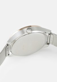 Reloj de acero inoxidable plateado con una caja redonda, acentos en tono dorado, correa de malla texturizada y logotipo de la marca grabado en la parte posterior.