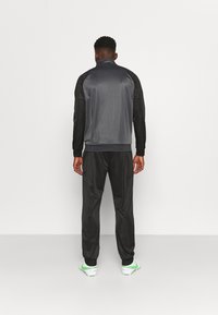 Trainingsjack en broek in grijs en zwart. De jack heeft een hoge kraag. De broek is taps toelopend, met elastische boorden en een gladde stof.