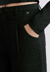 Zwarte, gestructureerde broek met een subtiel loofpatroon, voorzien van een zijzak en een zilveren logoknoop op de tailleband.