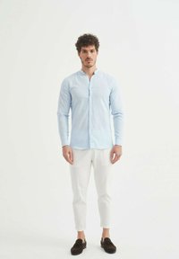 Chemise bleu clair à manches longues avec col mandarin, associée à un pantalon blanc fuselé et des mocassins marron foncé.