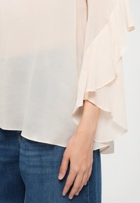 Ljusrosa, genomskinlig blus med volangärmar och rundad nederkant. I kombination med blå denimjeans som visar en lagerstruktur.