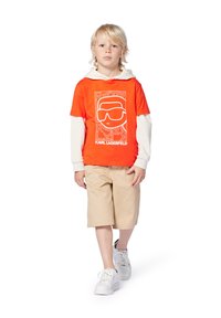 Oranje graphic t-shirt met witte print, laag over een witte longsleeve, gecombineerd met beige shorts en witte sneakers.
