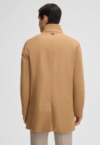 Cappotto in lana color cammello con vestibilità sartoriale, colletto alto e spacco posteriore singolo. Presenta una texture liscia e dettagli di design minimalisti.
