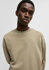 Felpa beige con collo a girocollo a coste e discreto logo HUGO. Il tessuto appare liscio e morbido, con una vestibilità comoda e spalle scese.