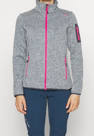 Person trägt eine graue Fleecejacke mit hell pinken Reißverschlüssen und blaue Hose vor einfachem weißem Hintergrund.