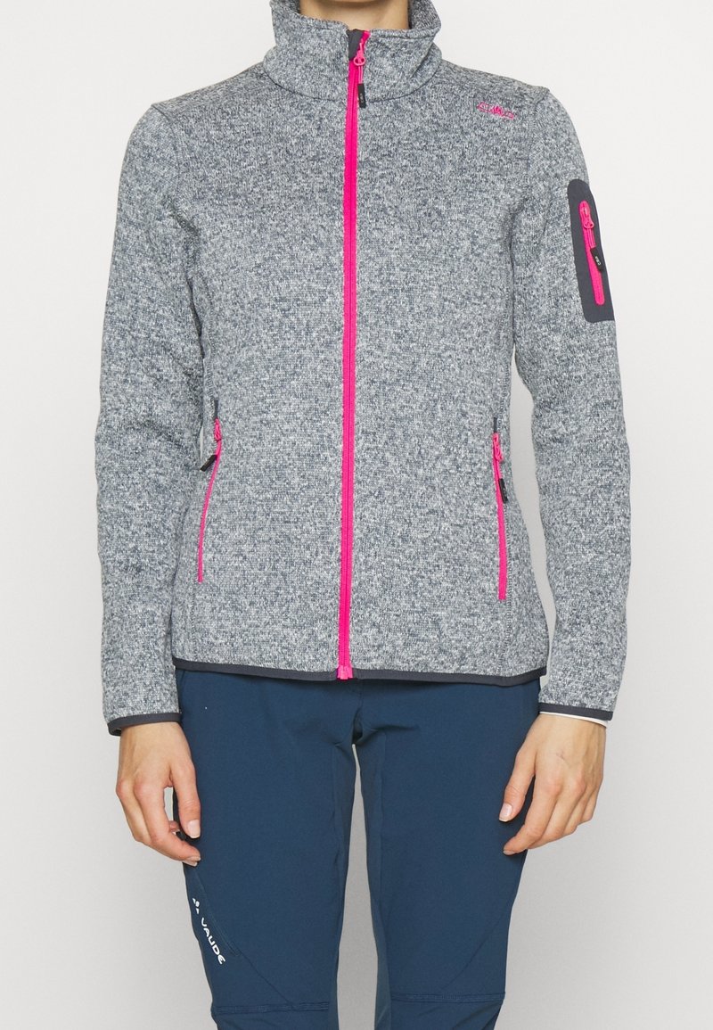 Personne portant une veste polaire grise à fermeture éclair rose vif et un pantalon bleu sur un fond blanc uni.
