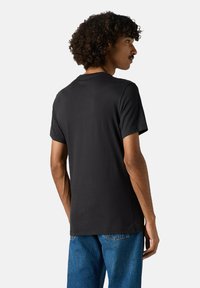 Mand med krøllet hår iført en tætsiddende ensfarvet sort t-shirt og blå jeans, der står i siden mod en lys grå baggrund.
