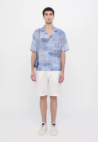 Chemise à manches courtes bleue à motifs avec un short blanc. La chemise présente diverses nuances et textures, associée à des baskets blanches et un sac bleu.