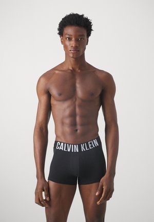 Calzoncillos boxer negros con una ancha cinturilla blanca que presenta el logo "CALVIN KLEIN"; material de algodón suave, diseño ajustado, longitud hasta medio muslo.
