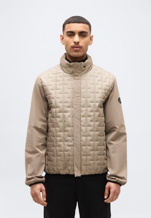 Άνδρας με κοντά μαλλιά που φοράει μπεζ μπουφάν με ύφανση και ψηλό γιακά και μαύρο παντελόνι, στέκεται μπροστά σε απλό λευκό φόντο.