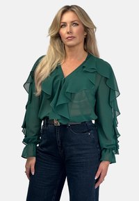 Green sheer blouse with ruffle details, v-neckline, and long sleeves; paired with dark denim high-waisted jeans.