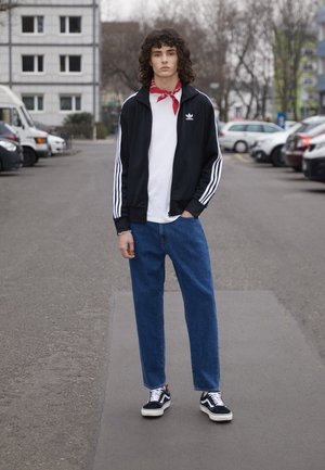 Черен Adidas анцуг с бели ивици, бяла тениска, сини широки дънки и черни кецове Vans. Червена кърпа около врата.