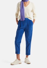 Pantalones de corte ancho de pana azul con frente plisado, combinados con un suéter de cuello en V color crema y una bufanda de punto morada. Zapatillas blancas.