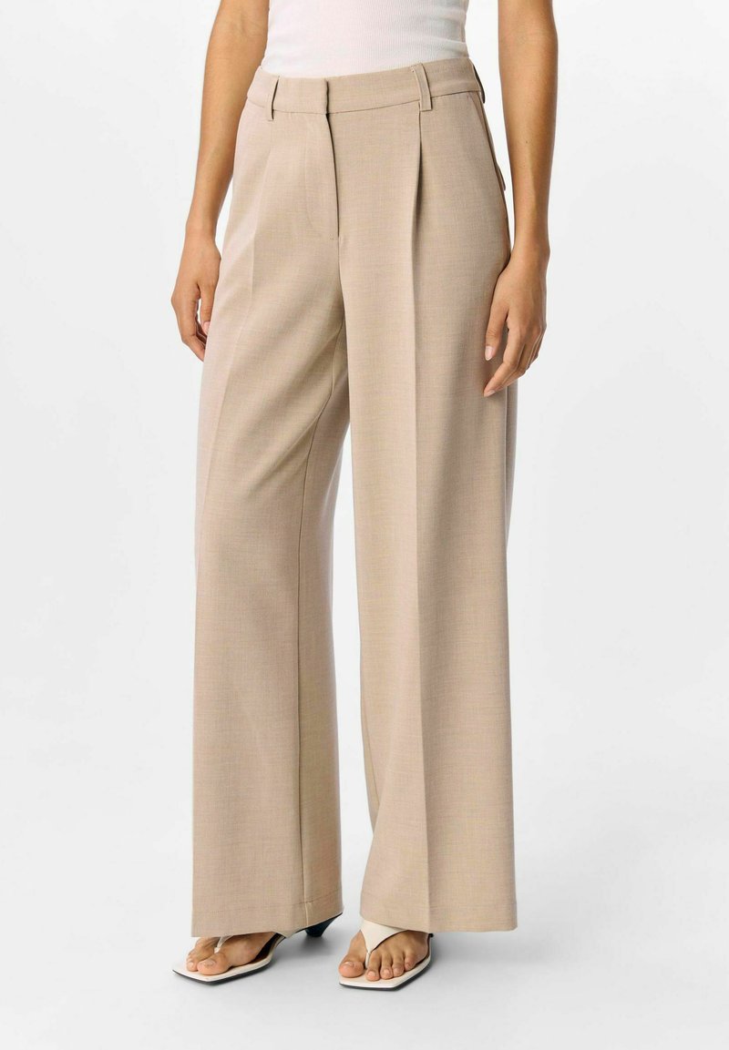 Beige wijde pijpen broek met plissee, gemaakt van een textuurstof. Beschikt over een hoge taille en subtiele zijzakken, gecombineerd met witte sandalen.