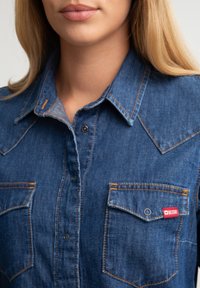 Camisa de mezclilla en azul medio con botones a presión, cuello puntiagudo y dos bolsillos en el pecho. Presenta costuras en contraste y una etiqueta de marca.