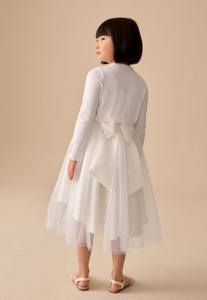 Pige med kort sort hår iført en hvid kjole med tylskørt, cardigan og stor sløjfe bagpå, stående mod en beige baggrund.