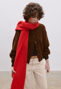 Hnědý pletený kardigan s velkými knoflíky, doplněný jasně červenou texturovanou šálou. Světlé kalhoty s širokými nohavicemi dokončují outfit.