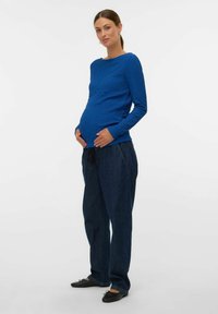 Top lungo blu con texture e maniche lunghe, dotato di scollatura rotonda, abbinato a pantaloni di denim scuri e ballerine nere. Il top si adatta in modo largo su un pancione visibile.