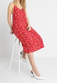 Robe maxi à motifs floraux rouges avec des fines bretelles, offrant une coupe décontractée et en tissu léger. Des chaussures à talons carrés blanches complètent le look.