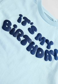 Helles blaues T-Shirt mit großen, dunkelblauen, puffigen Buchstaben, die "Ich habe Geburtstag" sagen. Glatte, weiche Textur und gerippter Halsauschnitt.