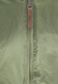 Olive green jacket featuring a central zipper with gold teeth and a brown leather pull tab, smooth texture, and clean stitching.