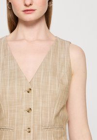 Femme portant un gilet beige sans manches à col en V avec des rayures blanches verticales et trois boutons, montrant la partie inférieure du visage et le cou.