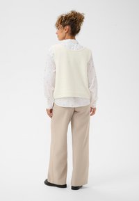 Kremowa dzianinowa kamizelka na białej koszuli z kołnierzykiem i subtelnym wzorem w kropeczki, zestawiona z beżowymi szerokimi spodniami i czarnymi butami.