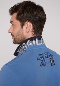 Niebieska koszulka polo z kołnierzykiem stójkowym z wzorzystymi granatowymi detalami. Haftowane napisy i logo w ciemniejszym odcieniu niebieskiego na plecach.