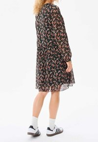 Nainen, jolla on päällään musta kukallinen pitkähihainen mekko, valkoiset sukat ja valkoiset tennarit mustilla raitoja, seisoo yksinkertaista valkoista taustaa vasten.