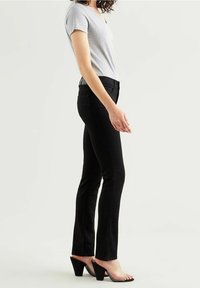 Femme portant un pantalon noir ajusté, un haut gris clair à manches courtes et des sandales à talons noirs avec des lanières transparentes, debout de profil.