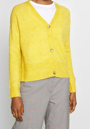 Cardigan jaune boutonné porté sur une chemise blanche, associé à un pantalon à carreaux noir et blanc avec de fines lignes rouges.
