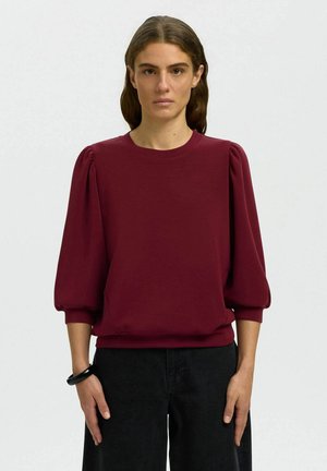Femme aux cheveux bruns ondulés portant un haut bordeaux à manches bouffantes et un pantalon noir, debout devant un fond clair uni.