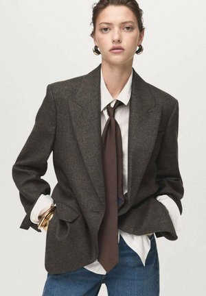 Blazer à carreaux marron avec larges revers, associé à une chemise blanche à rayures et une cravate marron foncé. Accentué par des bracelets jonc en or.