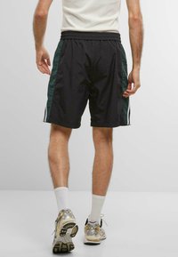Sorte træningsshorts med grønne sidelister, elastisk talje og hvide accentstriber. Bærer en grå top og løbesko.