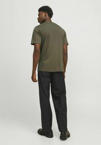 Temno olivnozeleni kratkosleečni T-shirt z okroglo ovratnikom, v kombinaciji s črnimi hlačami in črnimi čevlji, viden odzadaj.