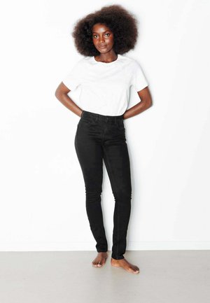 Femme aux cheveux afro debout pieds nus contre un mur blanc, portant un t-shirt blanc et un jean noir ajusté.