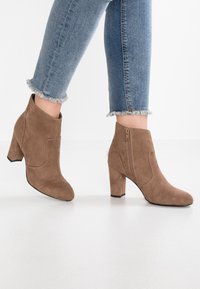 Hnědé semišové kotníkové boty s robustním podpatkem, bočním zipem a prošitými detaily. Nošené s modrými džínami s opraným lemem. Hladká textura.