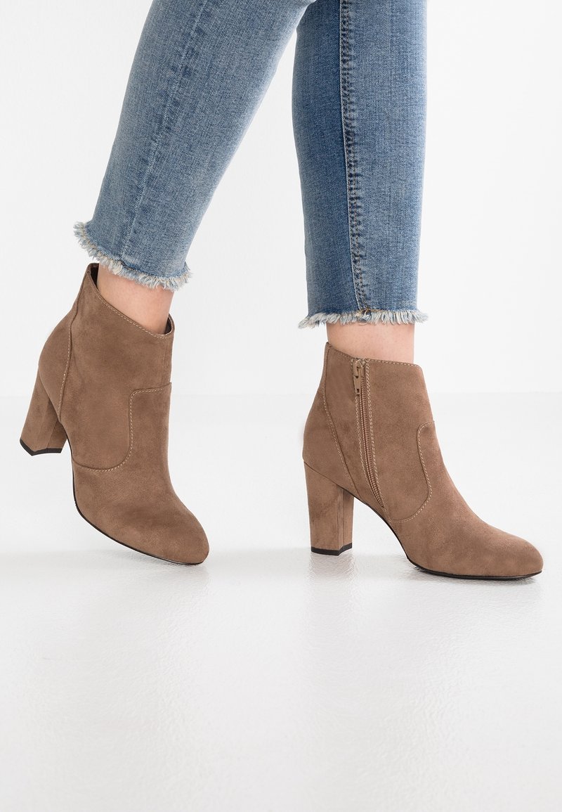 Hnědé semišové kotníkové boty s robustním podpatkem, bočním zipem a prošitými detaily. Nošené s modrými džínami s opraným lemem. Hladká textura.