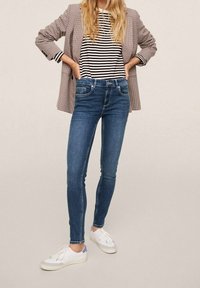 Jeans ajustados azules, camiseta de rayas en blanco y negro, blazer de cuadros grises y zapatillas blancas. Las características incluyen un diseño ceñido y una textura clásica de mezclilla.
