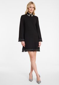 Robe noire avec un col et une bordure semblable à des perles. Manches longues avec des poignets et un ourlet froncés. Matériel lisse, longueur au genou.